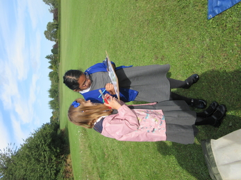 Two schoolchildren stand in a field looking at a clipboard together, with one holding a bug pot