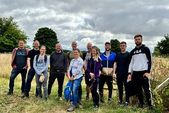 A group of 11 Midwich staff stand in a field on a grey day and smile at the camera, they are dressed in casual clothing