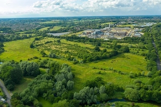 An aerial view over Sweet Briar Marshes. There is a large green space, broken up by patches of green trees and bushes, with houses and business buildings in the distance, all under a blue sky with some fluffy white clouds.