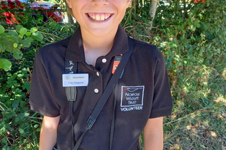 A photo of Toby smiling and wearing his NWT t-shirt, cap and name badge. He is also carrying a pair of binoculars and a camera. 