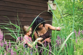 A child and a young person stand among purple wildflowers and point to them. The child has chin length dark blond hair and wears a pink t-shirt, while the young person has black curly hair and wears a brown vest top.