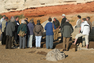 A group of people pictured from behind, gathered at the foot of a cliff. They are standing on sand in front of the orange and yellow cliff wall.