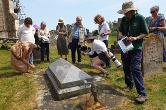 A group of people stand around a gravestone at Wymondham Abbey, looking down at the ground. There is a glass box containing a stuffed bird beside the gravestone.
