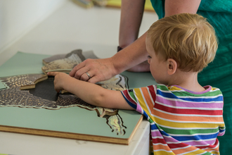 A young boy wearing a rainbow striped t-shirt completes a fossil puzzle, with a parent helping from behind
