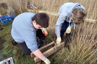 Two teenagers wearing grey hoodies build a wooden bridge on a grassy patch