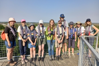 A group of young people standing in a row and smiling at the camera, while they stand on a raised platform above the trees on a sunny day