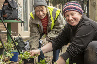 Two smiley community members are watering and potting small plants with big smiles on their faces.