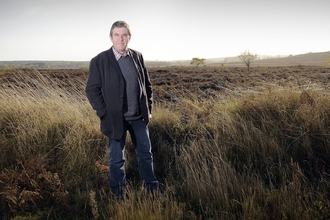 A man stands in a field of long grass and looks at the camera