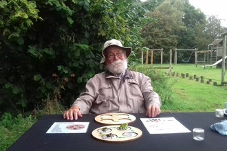 A volunteer sits at a table in a grassy field. He has a thick white beard and is wearing a light brown jacket and hat