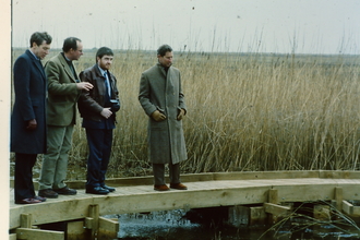 Prince Charles and three other men stand on a boardwalk with reeds behind them, as they look across a stretch of water
