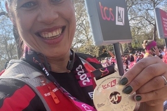 A woman wearing sports clothes holds a medal to the camera and smiles, in front of a crowd of runners