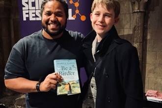 A man with black hair and a beard holds a book called 'Be a Birder'. He stands beside a teenage boy with blond hair. They are both smiling at the camera, in front of a poster for Norwich Science Festival.