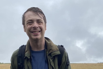 Alfie Bowen outdoors on a cloudy grey day, smiling at the camera, with backpack straps on his shoulders