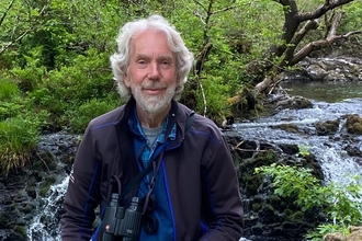 David North posing with his binoculars in front of a small waterfall. He has wavy grey hair and a grey beard.