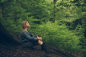 A woman sitting in woodland, with her arms drawn around her knees as she looks up into the distance