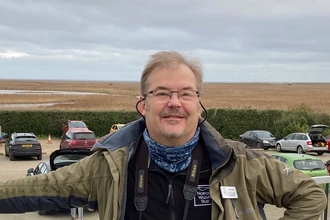 Tim is posing at Cley visitor centre in front of the car park, with his right arm outstretched and his left arm on his hip