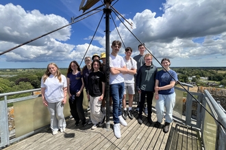 A group of young people on work experience standing together at the top of a tower on a sunny day, with the group all smiling at the camera