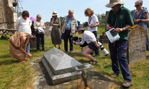 A group of people stand around a gravestone at Wymondham Abbey, looking down at the ground. There is a glass box containing a stuffed bird beside the gravestone.