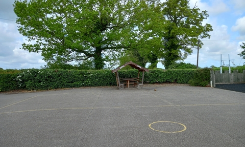 A school playground with a sheltered bench at the back, with trees behind the playground hedge