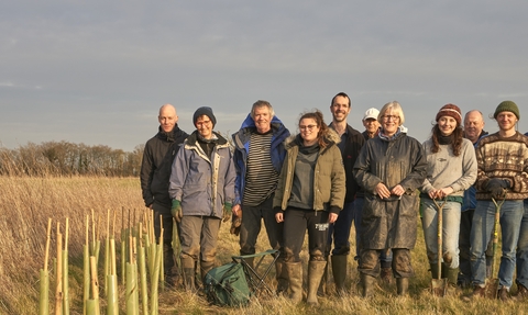 A group of people wearing muddy clothes and holding spades, who are standing in a field next to sticks marking newly planted trees