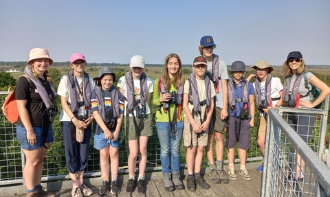 A group of young people standing in a row and smiling at the camera, while they stand on a raised platform above the trees on a sunny day