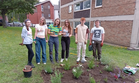 A group of teenagers standing in a line and smiling at the camera in front of a recently planted flower bed by a block of flats