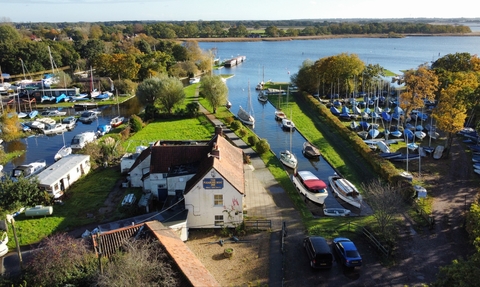 An aerial view on a sunny day of a pub building, which is next to a stretch of Broad filled with boats