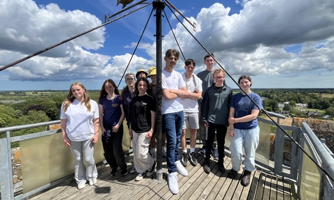 A group of young people on work experience standing together at the top of a tower on a sunny day, with the group all smiling at the camera