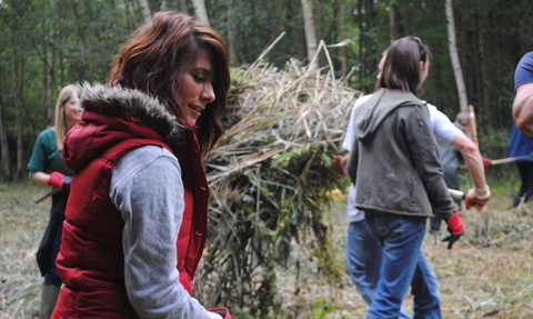 A woman with dark hair wearing a red gilet rakes up hay, while other volunteers work behind her
