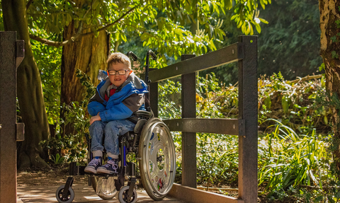 A young boy wearing glasses is grinning in his wheelchair whilst on a wooden bridge at a nature reserve
