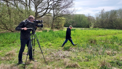A man stands behind a professional camera on a tripod in a field, while another man walks in front of the camera