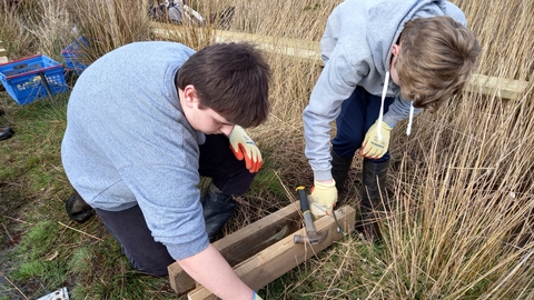 Two teenagers wearing grey hoodies build a wooden bridge on a grassy patch