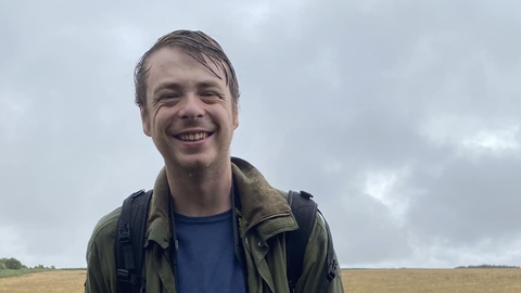 Alfie Bowen outdoors on a cloudy grey day, smiling at the camera, with backpack straps on his shoulders