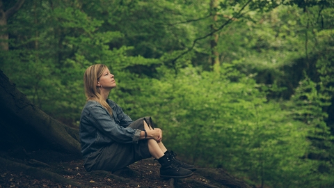 A woman sitting in woodland, with her arms drawn around her knees as she looks up into the distance