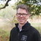 Norfolk Wildlife Trust CEO, Eliot Lyne, smiles at the camera while standing in front of woodland. He is wearing glasses and a black NWT fleece jacket