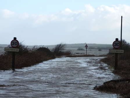 A flooded road at NWT Cley Marshes. The road is completely covered by water, with signs showing 30mph and Salthouse on each side of the road. Hills and trees can be seen in the distance.