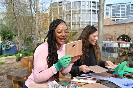 Two young people sit together smiling and building bird boxes in a garden by a canal. The person on the left has long black hair in braids and is wearing a pink jumper and green gloves. The person on the right has long curly dark brown hair and is wearing a black jumper.