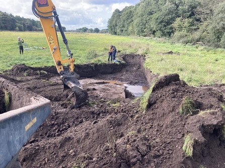 A digger works on excavating a ghost pingo. It digs up earth, with some water visible at the bottom of the earth. Three people are visible standing on the grass by the pond which is being excavated.