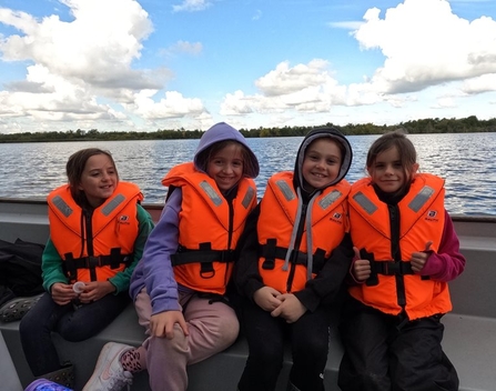 Four children from Acle Primary School sit on a boat at NWT Ranworth Broad on a sunny day, all wearing bright orange high-vis lifejackets and smiling at the camera