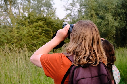 Two people pictured from behind, using binoculars to look up into a tree at Sweet Briar Marshes. The person in the foreground has shoulder length dark blonde hair and is wearing an orange t-shirt and purple backpack.