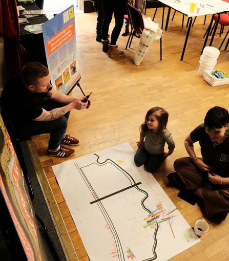 A child sits on the wooden floor of a hall beside an adult, while another adult sits on a ledge beside them. They are sitting in front of a large sheet of paper, which has coloured pens on. The paper has been drawn on, with ideas for the nature reserve.