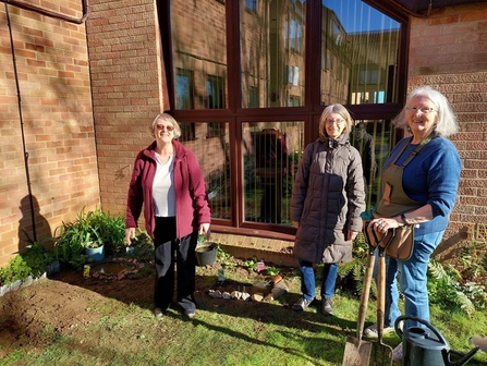 Three people stand in front of a building on a sunny day, holding gardening equipment and smiling at the camera. They are all wearing winter coats and have grey, shoulder length hair