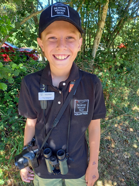 A photo of Toby smiling and wearing his NWT t-shirt, cap and name badge. He is also carrying a pair of binoculars and a camera. 