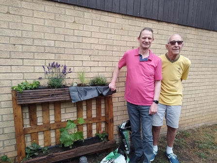 Two people stand beside a wooden pallet planter, filled with green plants and lavender. One of the people wears a pink t-shirt and blue jeans, while the other wears a yellow t-shirt and grey shorts and sunglasses. Both are smiling at the camera