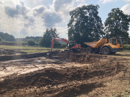 A yellow excavator digs a large hole beside a yellow dump truck in a grassy field. The hole is full of brown mud and is dug in a square shape.