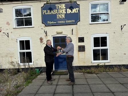 Two men stand in front of the entrance to a pub, which features a sign saying 'The Pleasure Boat Inn'. The men are smiling and looking at each other, while they shake hands with their right hands and exchange a set of keys with their left hands.