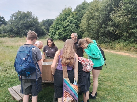 A group of teenagers stand around a wooden box examining its contents, while standing in a field