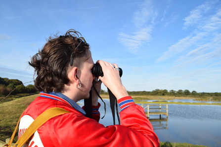 A young person with shoulder length brown hair and an earring in their right ear, wearing a red jacket, looks through binoculars at an area of marsh