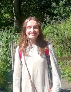 A young person with shoulder length brown hair looks at the camera and smiles in front of greenery