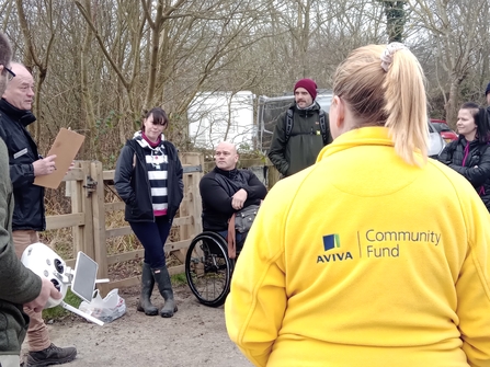A group of people stand in a circle listening to instructions, with a blonde woman wearing a yellow Aviva Community Fund fleece in the foreground